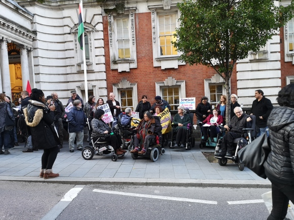 Group of people on a pavement, some in wheelchairs, some carrying banners and placards