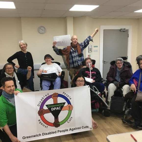 9 people in group photo, 5 wheelchair users, 6 women, 3 men, all Caucasian one East Asian, 2 kneeling up front holding banner 'Greenwich Disabled People Against Cuts ' logo